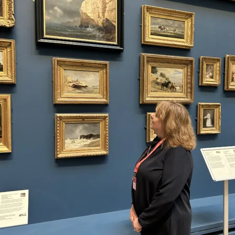 A woman looking at paintings in a gallery. The gallery has dark blue walls. She is focused on a frame that is black, with a painting of a seascape.