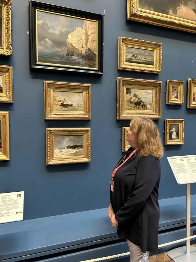A woman looking at paintings in a gallery. The gallery has dark blue walls. She is focused on a frame that is black, with a painting of a seascape.