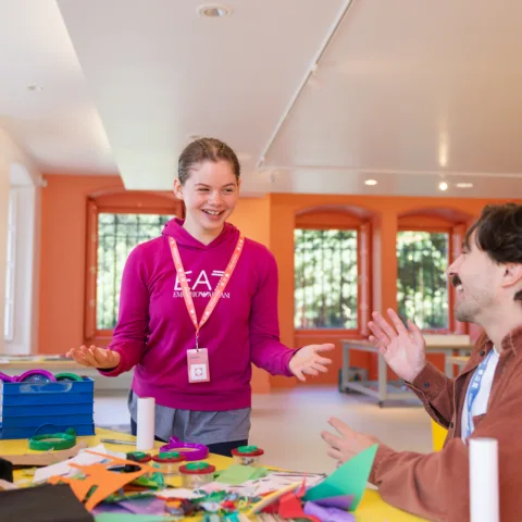 A girl and a man smile and talk at a craft table covered with colorful paper, scissors, and other art supplies in a bright room.