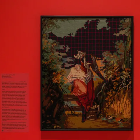 Framed needlework artwork titled 'Study in Pink and Gray by Matt Smith' is displayed against a vivid pink wall, featuring a classical pastoral scene with a pixelated grid obscuring the upper portion.