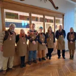 Seven volunteers wearing brown aprons and name badges stand smiling in front of a glass-fronted bookcase filled with colourful, neatly arranged books in a warmly lit room with wooden floors.