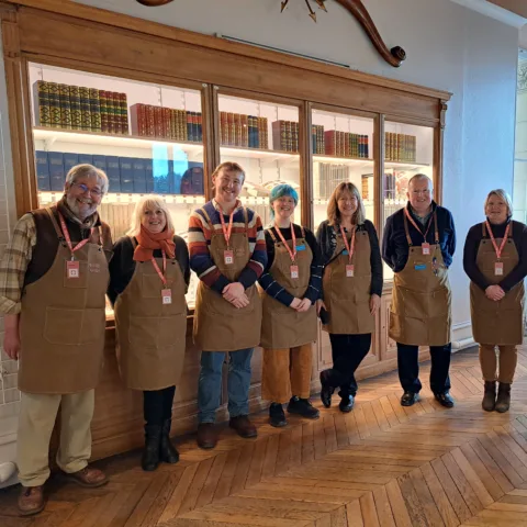 Seven volunteers wearing brown aprons and name badges stand smiling in front of a glass-fronted bookcase filled with colourful, neatly arranged books in a warmly lit room with wooden floors.