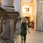 A young child with blonde hair and wearing green dungarees walks along a long corridor of a museum, surrounded by large stone pillars and large arching doorways.