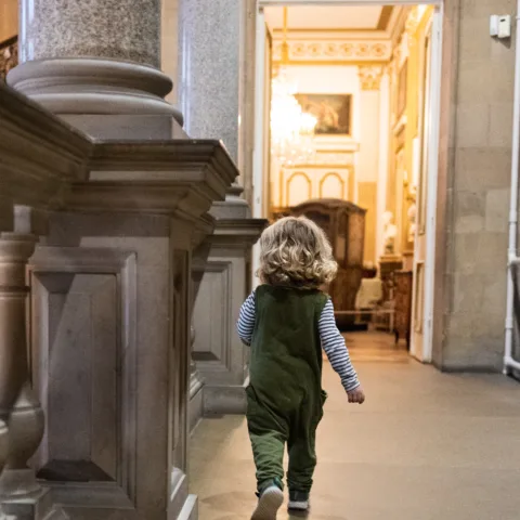 A young child with blonde hair and wearing green dungarees walks along a long corridor of a museum, surrounded by large stone pillars and large arching doorways.