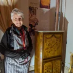 A smiling woman with short grey hair stands beside an ornate wooden cabinet decorated with floral marquetry. She wears a striped apron over a black outfit, black gloves, and a red Bowes Museum lanyard. Behind her is a display panel titled "Baroque 1685 to 1725," describing the opulent style. The scene is warmly lit, highlighting the craftsmanship of the furniture.