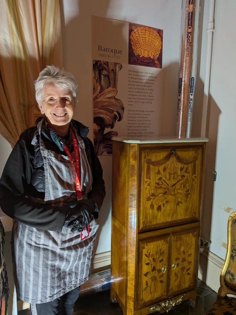 A smiling woman with short grey hair stands beside an ornate wooden cabinet decorated with floral marquetry. She wears a striped apron over a black outfit, black gloves, and a red Bowes Museum lanyard. Behind her is a display panel titled "Baroque 1685 to 1725," describing the opulent style. The scene is warmly lit, highlighting the craftsmanship of the furniture.