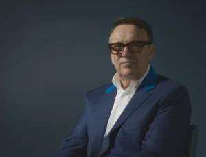 A man wearing a blue pinstripe suit, white shirt, and glasses sits on a wooden chair against a dark blue background, posed with hands clasped.