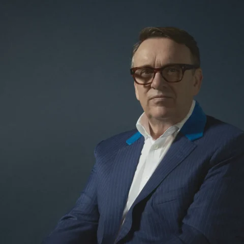 A man wearing a blue pinstripe suit, white shirt, and glasses sits on a wooden chair against a dark blue background, posed with hands clasped.