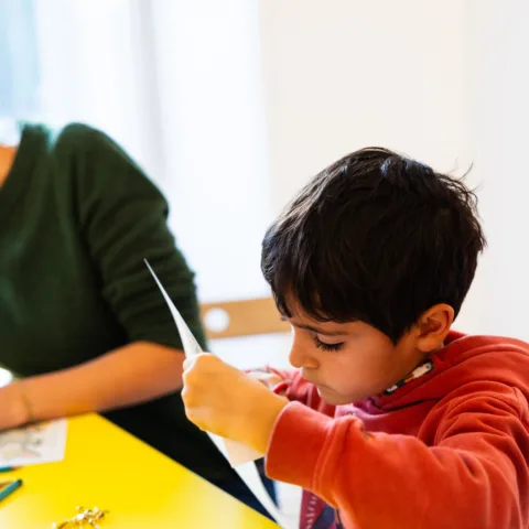 A young boy wearing a red hoodie is intently cutting a piece of paper with scissors while seated at a bright yellow table. His expression is focused and absorbed in the creative task. An adult, partially out of frame, sits beside him in a green jumper.