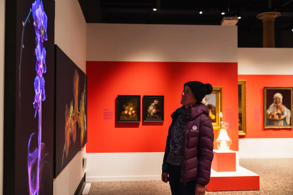A woman in a purple puffer jacket and black knit hat stands inside an art gallery, looking at a display of vibrant, illuminated floral artwork on the left wall. The gallery features pink accent walls and a variety of classical paintings, including still lifes and a portrait of a woman holding a basket of fruit.