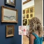 A woman with curly blonde hair wearing a teal sleeveless top looking at a museum guide in front of a deep blue wall displaying three framed paintings, with a bright, art-filled gallery visible through an arched doorway.