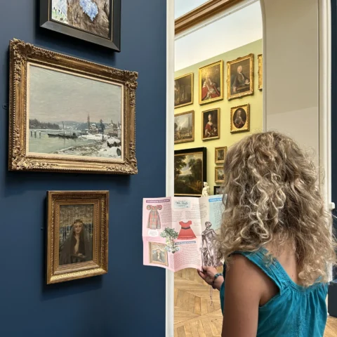 A woman with curly blonde hair wearing a teal sleeveless top looking at a museum guide in front of a deep blue wall displaying three framed paintings, with a bright, art-filled gallery visible through an arched doorway.