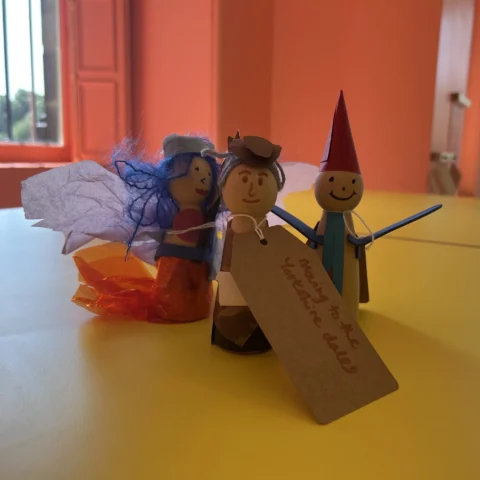 Three colourful peg dolls, dressed as whimsical characters with handmade accessories, stand on a yellow table in a brightly lit pink room, with a tag reading 'Moving to the Yorkshire Dales'.