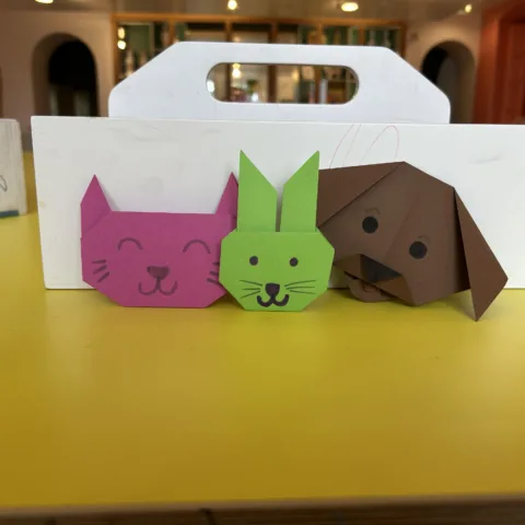 Three colourful origami animals—a pink cat, green rabbit, and brown dog—displayed on a yellow table in a creative workshop setting.
