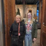 Two smiling women wearing red Bowes Museum lanyards stand in a grand doorway. The woman on the left has short blonde hair and is dressed in a long black padded vest with a crossbody bag. The woman on the right has long blonde hair and glasses, and is wearing a green parka over a festive jumper with reindeer and snowflake patterns, along with light blue jeans. Behind them, the Museum's ornate interior and a decorated Christmas tree are visible.