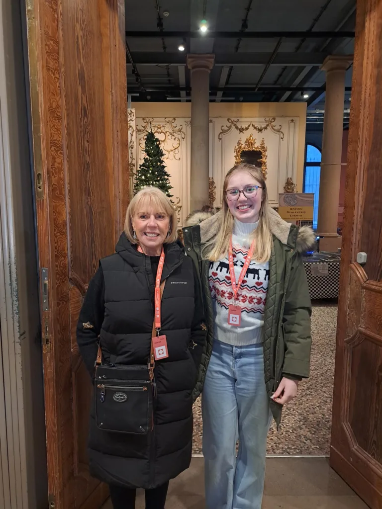 Two smiling women wearing red Bowes Museum lanyards stand in a grand doorway. The woman on the left has short blonde hair and is dressed in a long black padded vest with a crossbody bag. The woman on the right has long blonde hair and glasses, and is wearing a green parka over a festive jumper with reindeer and snowflake patterns, along with light blue jeans. Behind them, the Museum's ornate interior and a decorated Christmas tree are visible.