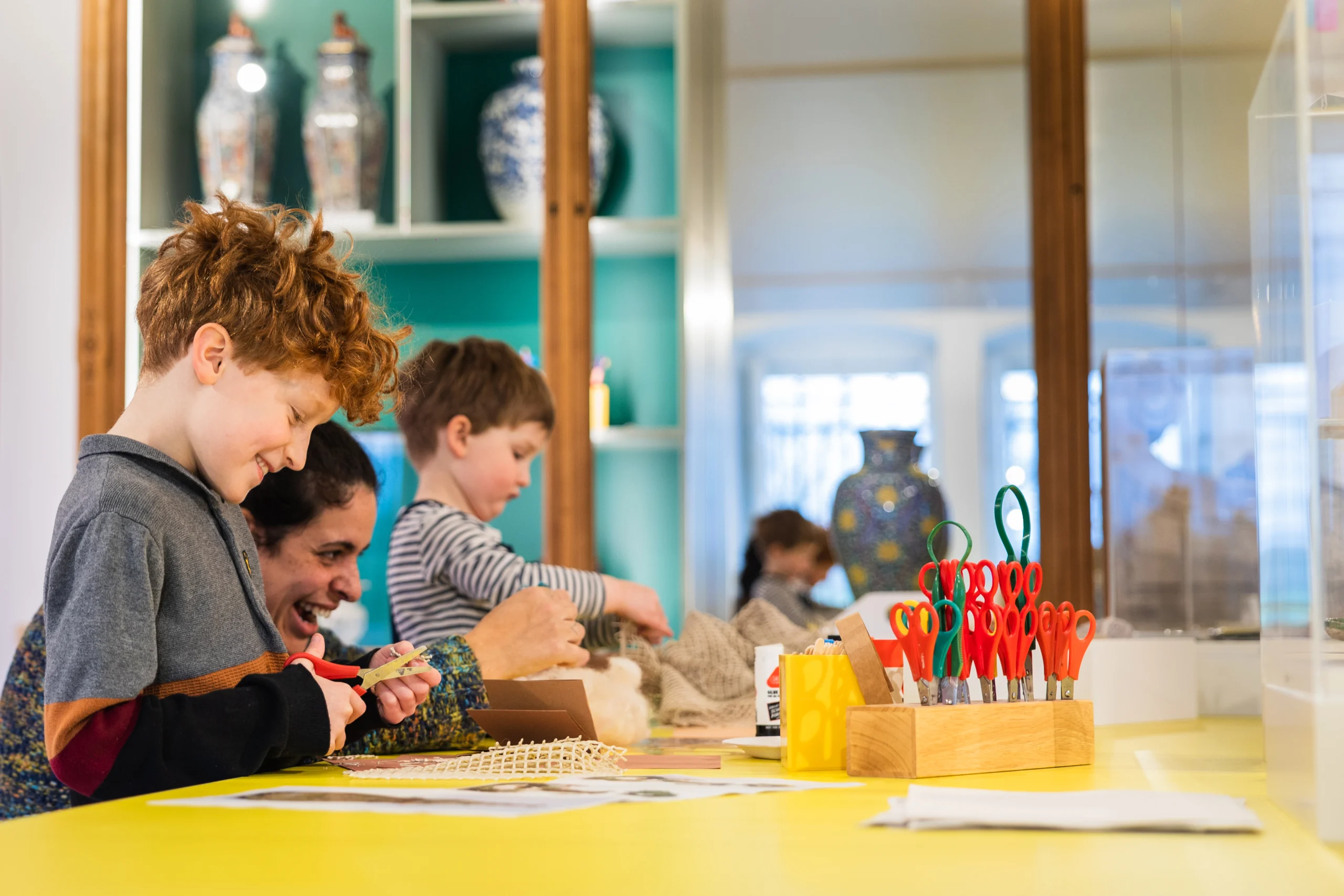 Two young children and an adult engage in an arts activity at a brightly lit table surrounded by colourful decorations and supplies including scissors, glue sticks and textured materials. There is a display of historical objects behind them in a museum display case.