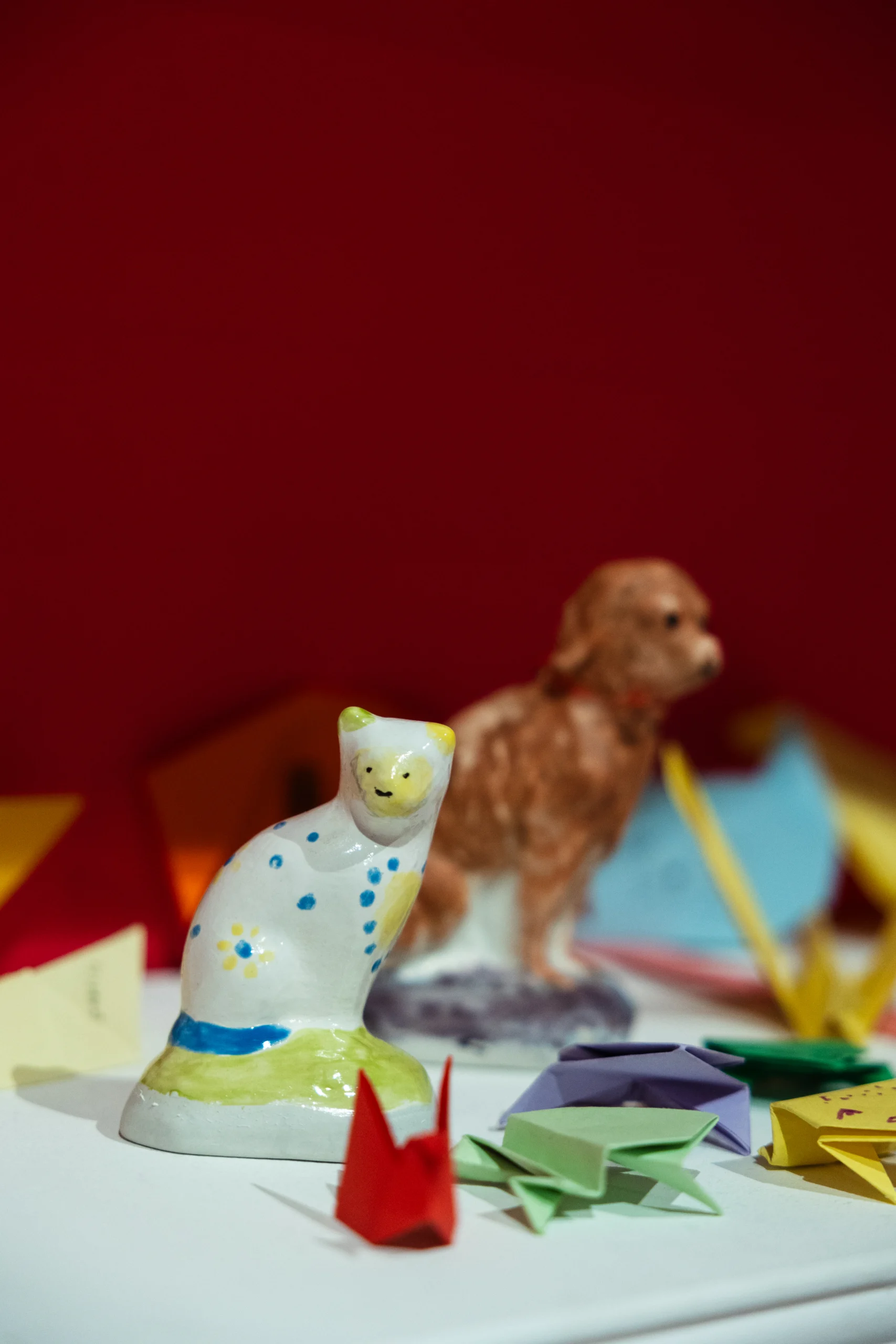 Close-up of a display featuring two small ceramic animal figurines—a white cat with blue and green painted details in the foreground and a brown dog positioned slightly behind. Surrounding the figurines are colorful origami pieces in red, green, purple, and yellow, set against a bright red background.