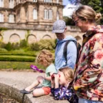 A family enjoys outdoor exploration in the gardens of The Bowes Museum, with two children using binoculars and a magnifying glass near the water’s edge.