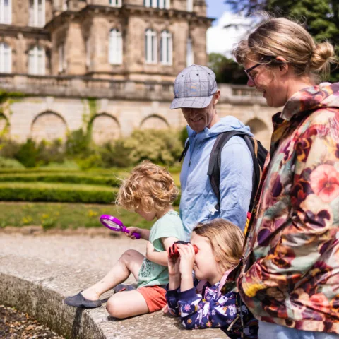 A family enjoys outdoor exploration in the gardens of The Bowes Museum, with two children using binoculars and a magnifying glass near the water’s edge.