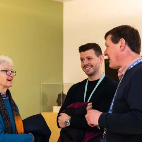 An older woman with curly white hair and glasses chats with two men wearing lanyards in a brightly lit gallery space. All three are smiling and appear engaged in conversation.