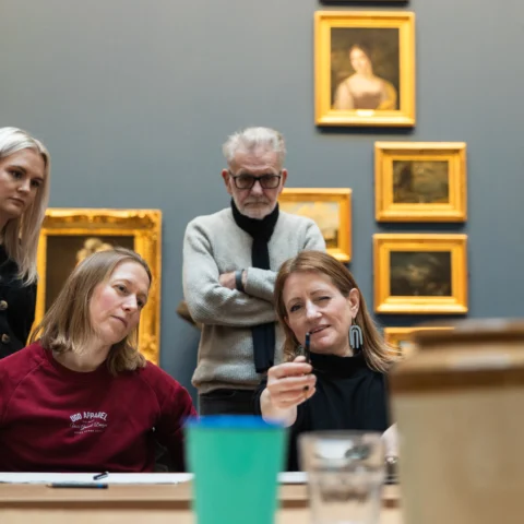 A group of five people, four women and one man, gathered around a table in an art gallery, closely examining an object in a woman's hand. Framed paintings hang on the wall behind them. The setting suggests a collaborative or educational environment, possibly an art workshop or discussion.