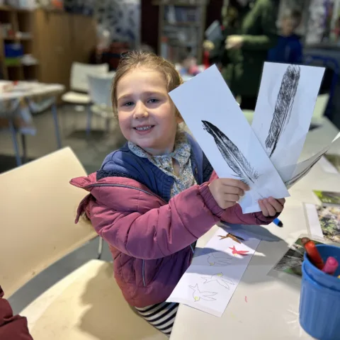 A smiling young girl in a pink coat proudly holds up two prints of feathers she made during a craft activity. She’s seated at a table covered in art supplies, with bird drawings and coloured pens visible. The background shows a creative space with shelves, books, and other participants.