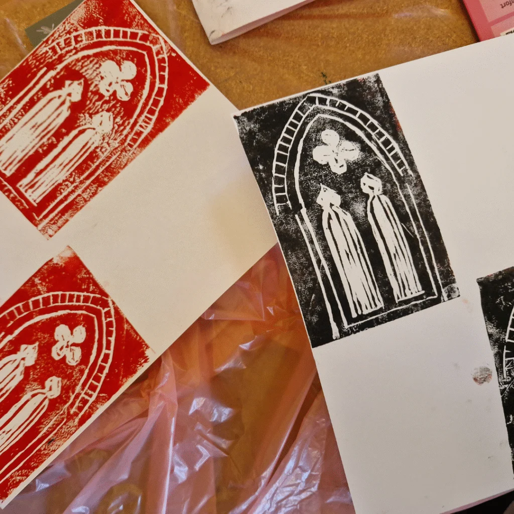 A set of block prints on white paper depicting two robed figures inside a pointed Gothic arch with a quatrefoil above. The prints are in red and black ink, arranged on a tabletop with art supplies partially visible.