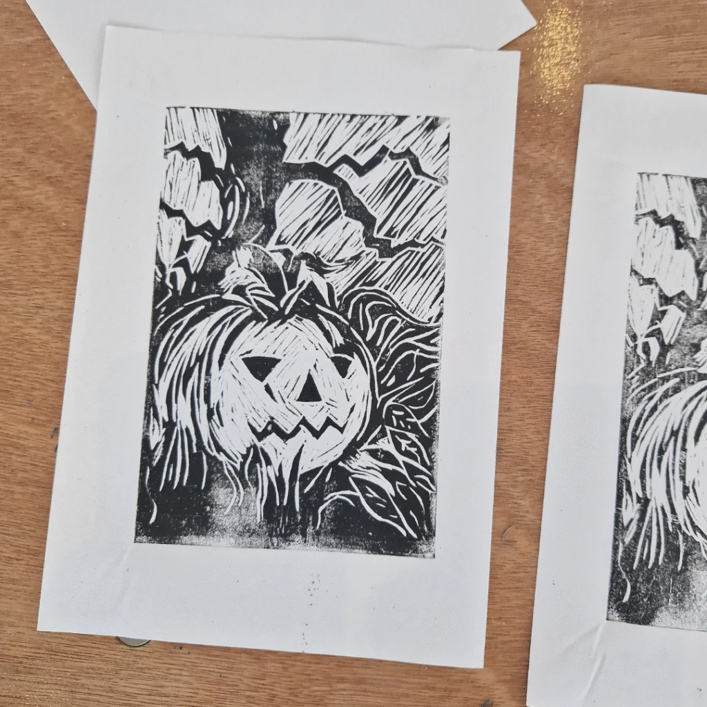 A black-and-white block print of a Halloween-themed design showing a carved jack-o’-lantern surrounded by curling vines and jagged shapes, printed on white paper.