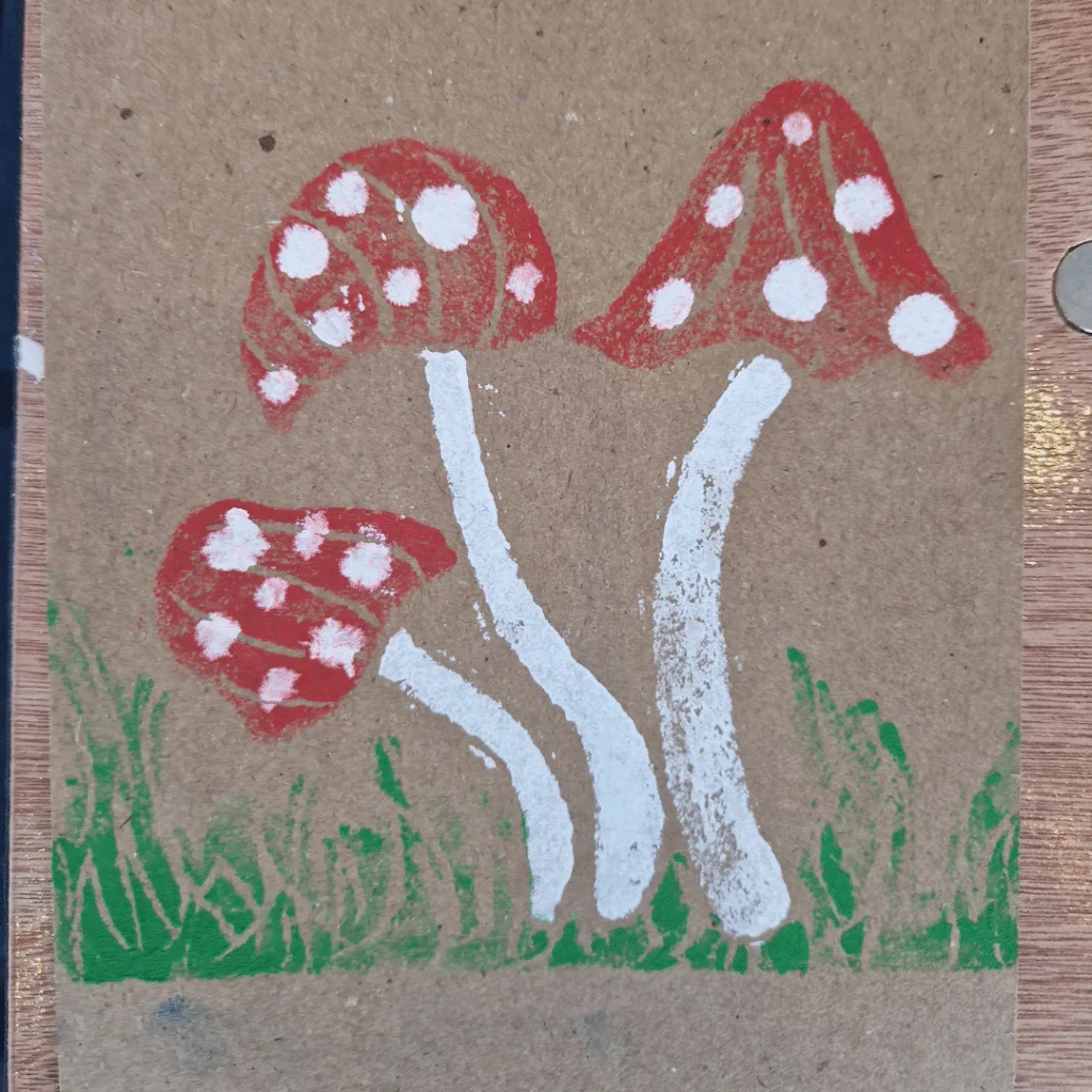 A block print of three red mushrooms with white spots and tall white stems, set against a brown background with green grass at the base.