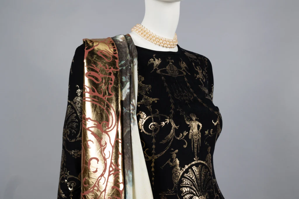 A mannequin dressed in an ornate black gown adorned with intricate gold classical motifs. Draped over one shoulder is a luxurious scarf or shawl with metallic gold designs on one side and multicolored patterns on the other, complemented by a pearl necklace.