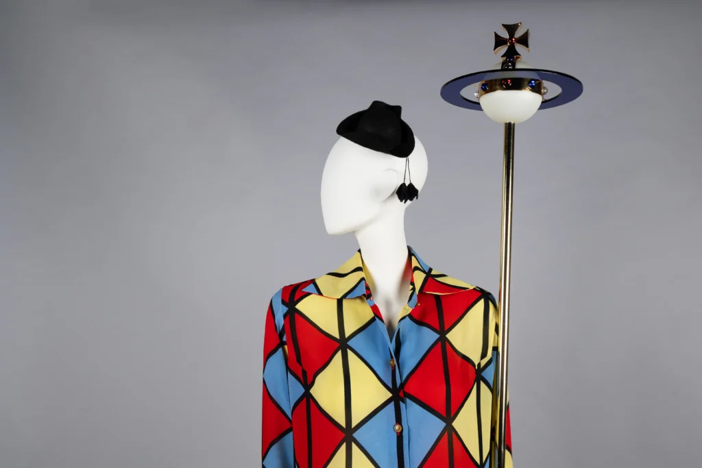 A mannequin wearing a vibrant jacket with a geometric diamond pattern in red, yellow, blue, and black. It is accessorized with a small black hat, a tassel-shaped earring, and stands beside a tall staff topped with an orb encircled by rings.