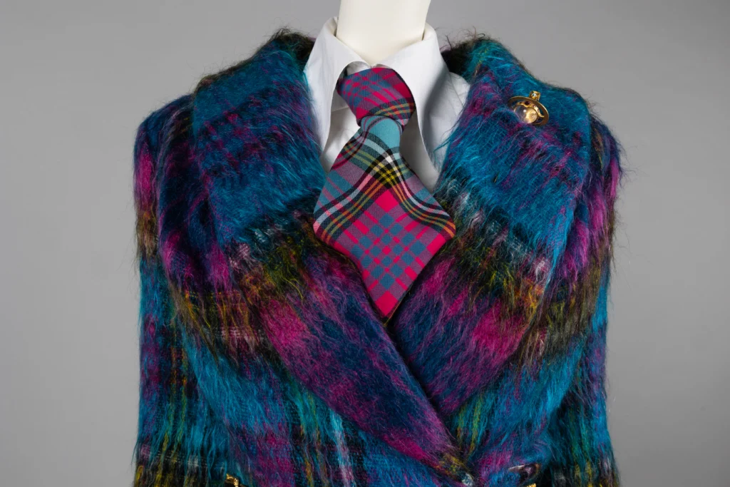 Close-up of a mannequin wearing a vibrant, multicoloured fuzzy coat in shades of blue, purple, and yellow. Underneath is a white shirt with a bold pink and teal tartan tie. A gold orb-shaped brooch is pinned to the coat’s lapel.