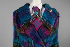 Close-up of a mannequin wearing a vibrant, multicoloured fuzzy coat in shades of blue, purple, and yellow. Underneath is a white shirt with a bold pink and teal tartan tie. A gold orb-shaped brooch is pinned to the coat’s lapel.