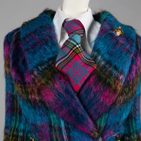 Close-up of a mannequin wearing a vibrant, multicoloured fuzzy coat in shades of blue, purple, and yellow. Underneath is a white shirt with a bold pink and teal tartan tie. A gold orb-shaped brooch is pinned to the coat’s lapel.