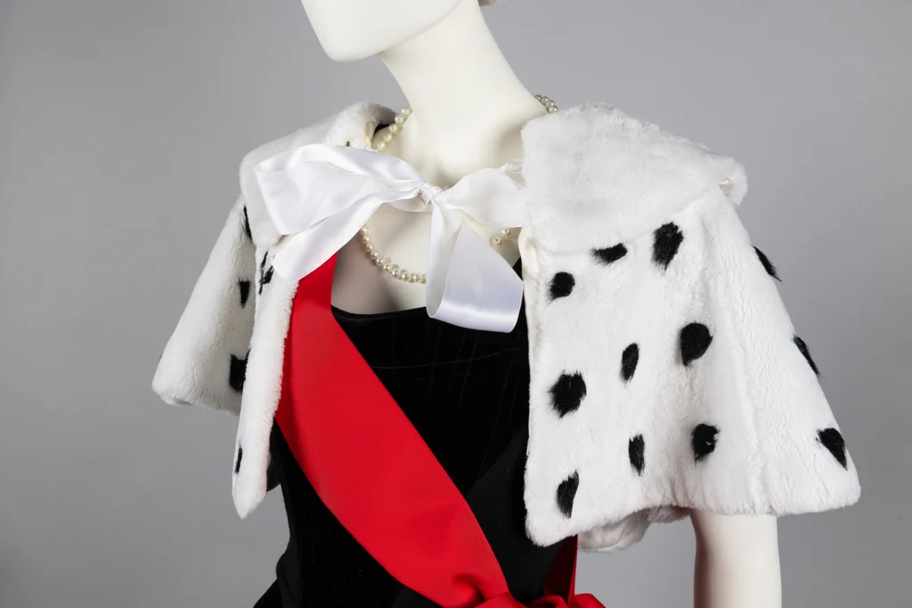 A mannequin styled in a sophisticated ensemble with a faux fur capelet featuring black spots, tied at the neck with a large white bow. Beneath the capelet is a black dress accented by a striking red sash across the chest.