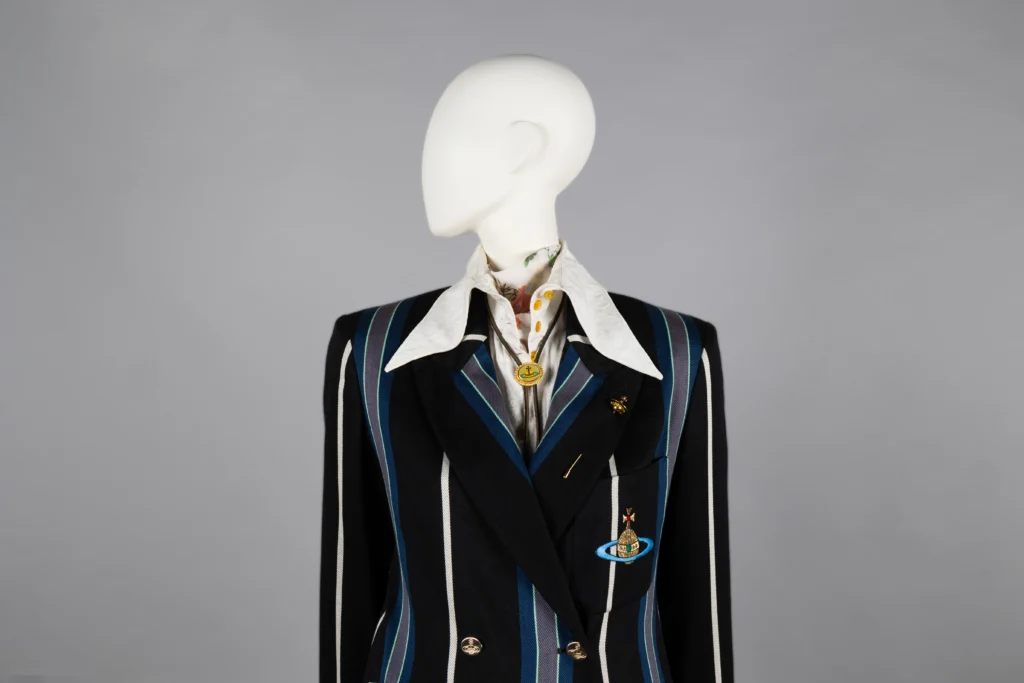 A mannequin dressed in a black double-breasted blazer with bold blue and white vertical stripes, featuring a large white collared shirt underneath. The blazer is embellished with gold buttons and two brooches—one shaped like a planet with rings and another circular pin.
