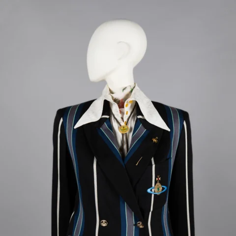 A mannequin dressed in a black double-breasted blazer with bold blue and white vertical stripes, featuring a large white collared shirt underneath. The blazer is embellished with gold buttons and two brooches—one shaped like a planet with rings and another circular pin.