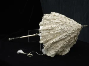 An ornate 19th-century lace parasol with an intricately patterned cream lace canopy. The handle is made of ivory or bone with a decorative metal finial and matching ring attachment, connected by a delicate tasselled cord. The parasol rests against a dark background, highlighting its fine craftsmanship and elaborate lacework.