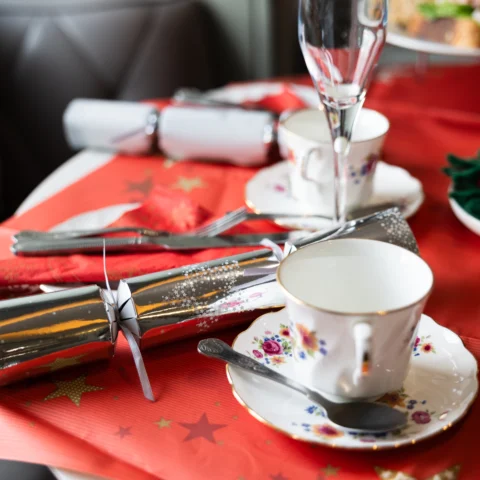 A festive table setting with fine china teacups and saucers decorated with floral patterns, placed on a red tablecloth with gold stars. Silver Christmas crackers, cutlery, and a champagne flute complete the arrangement. In the background, a tiered stand holds cakes and sandwiches, and a small green wreath with red berries adds to the Christmas theme.