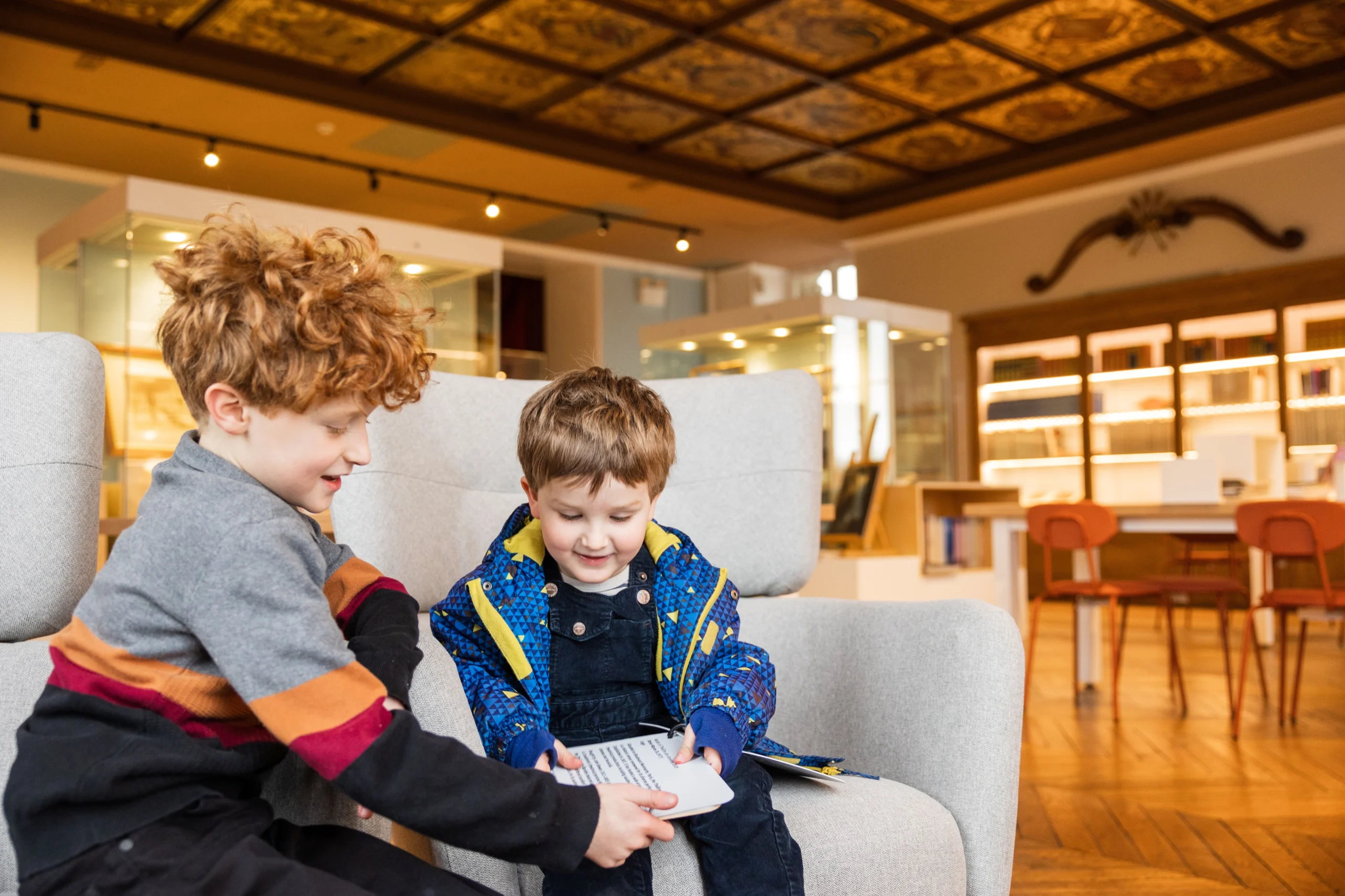 Two young boys sitting on a light grey armchair in a museum or library setting. The older boy, with curly red hair, is wearing a grey sweater with colored stripes and is showing a book to the younger boy, who is wearing a blue patterned jacket and smiling. In the background, display cases and bookshelves are visible, along with wooden flooring and a decorative ceiling.