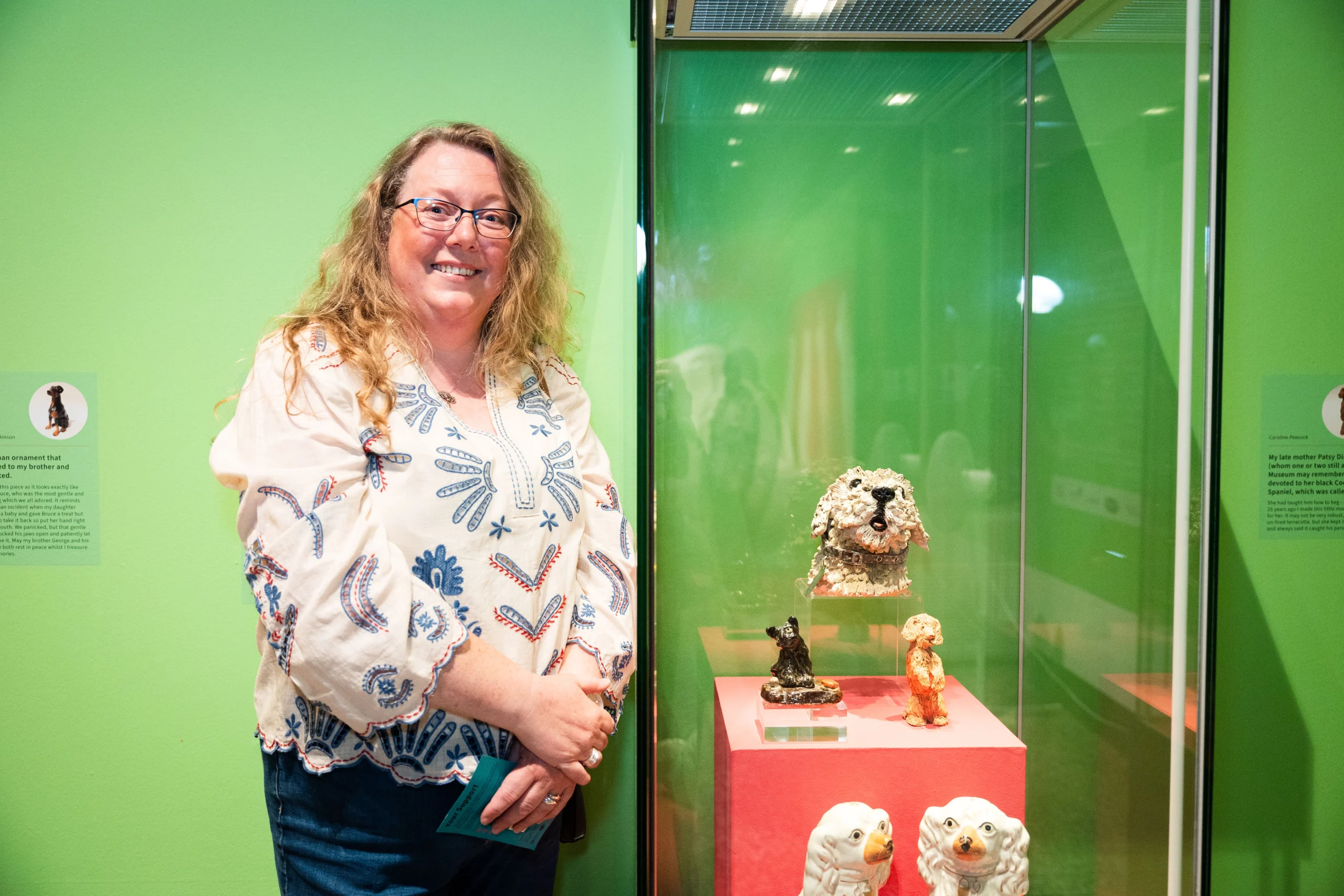 A woman with long blond hair and glasses stands smiling beside a glass display case in a museum. The display contains several ceramic dog figurines on a pink shelf. The background wall is bright green with descriptive text panels on either side.