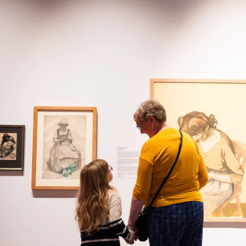An older woman in a yellow top and a young girl with long blonde hair stand hand in hand, looking at framed drawings and paintings of seated women displayed on a gallery wall. Warm lighting highlights the artworks and their quiet moment of connection.
