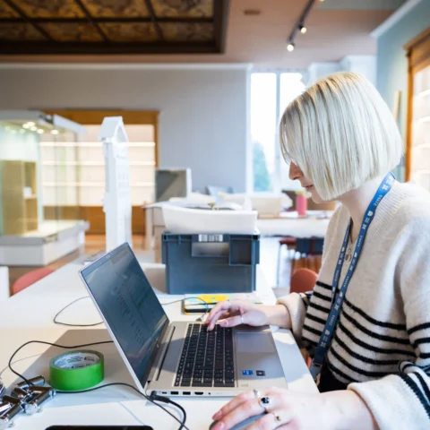 A person wearing a black and white striped sweater works on a laptop in a well-lit room with wooden cabinets and display shelves.