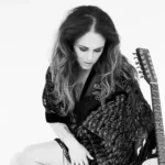 Black-and-white photo of a woman seated on a clear stool, looking down with loose wavy hair, wearing a patterned shawl and high-heeled boots, with an acoustic guitar resting beside her.