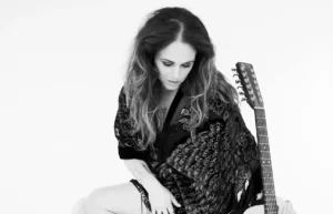 Black-and-white photo of a woman seated on a clear stool, looking down with loose wavy hair, wearing a patterned shawl and high-heeled boots, with an acoustic guitar resting beside her.