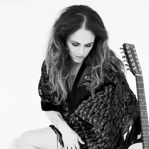Black-and-white photo of a woman seated on a clear stool, looking down with loose wavy hair, wearing a patterned shawl and high-heeled boots, with an acoustic guitar resting beside her.
