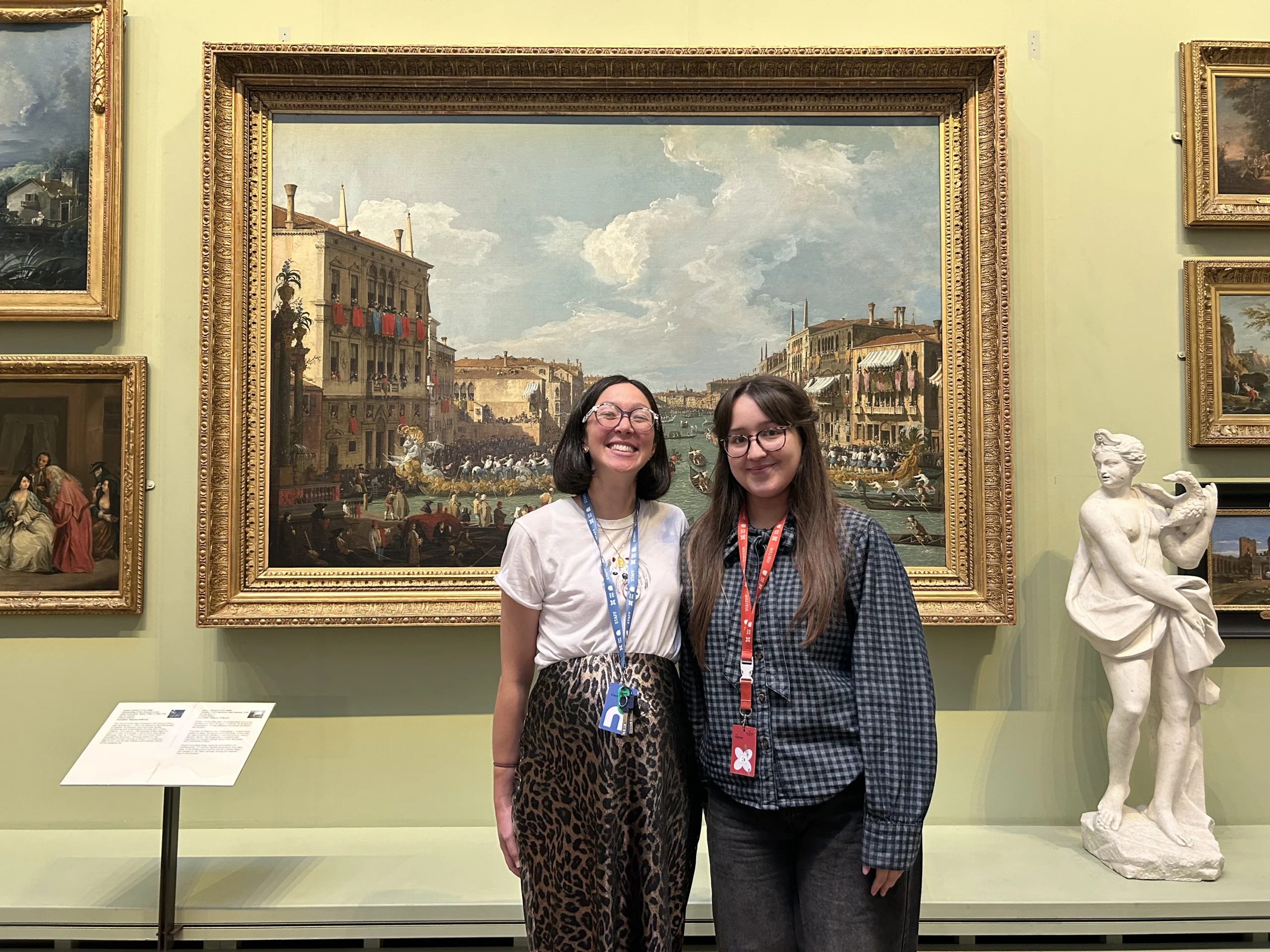 Two women standing and smiling in front of a large framed painting in an art gallery. One wears a white shirt and patterned pants, and the other wears a blue checked shirt and dark jeans. Several other framed artworks and a white statue are visible on the wall around them.