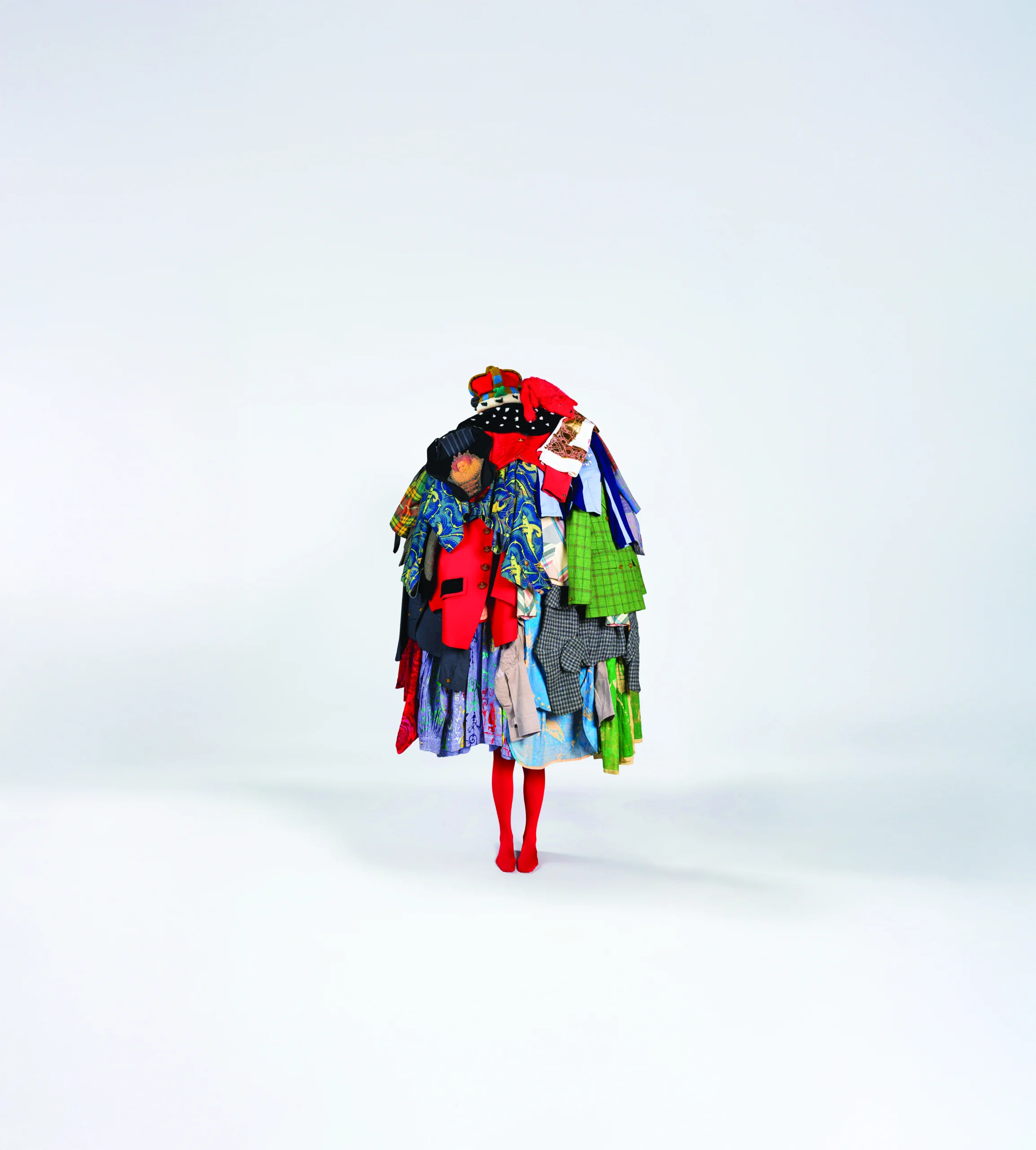 A full‑body figure stands against a plain light background wearing an elaborate sculptural outfit made from many layered garments. The clothing pieces are brightly coloured and vary in patterns and textures, including jackets, shirts, skirts, and fabric panels stacked and draped to form a voluminous, mound‑like silhouette. The figure also wears bright red tights and matching shoes, with only the legs visible beneath the mass of clothing.