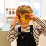 A young child wearing striped long sleeves and dark overalls smiles while holding up large, playful yellow sunglasses decorated with orange fuzzy eyebrows. The child stands indoors in a brightly lit room with soft-focus windows and displays in the background.